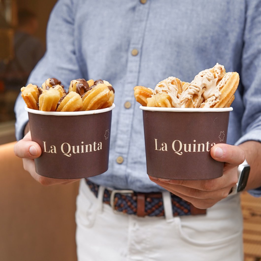 Two branded ice cream cups from La Quinta filled with churros, topped with chocolate and caramel drizzle.